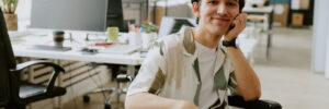 A man with brown hair sitting in his wheelchair looking at the camera and smiling. He is at work, with a desk and computer and shared office space in the background.